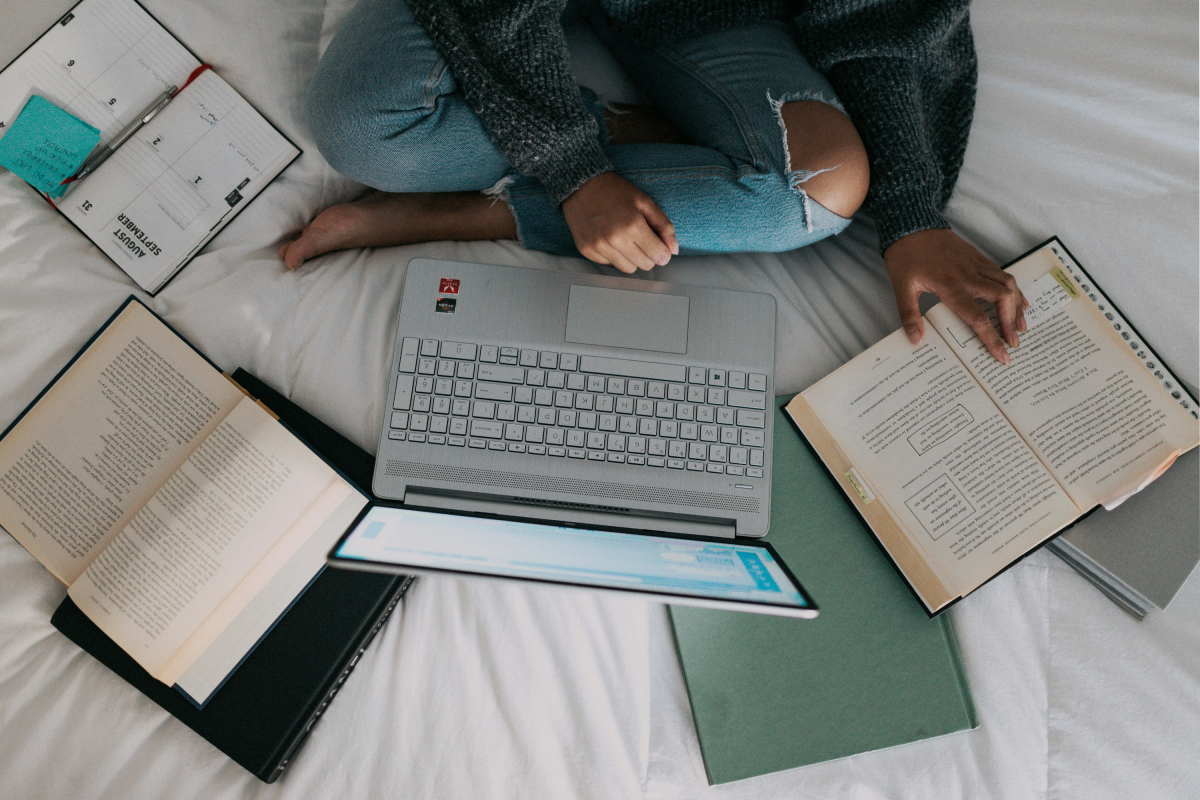 A student sat on their bed with a laptop and books for research.