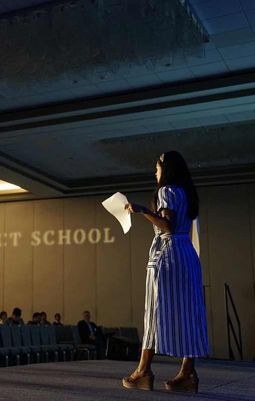 Student standing on stage with a paper in their hand, talking to a group of people.